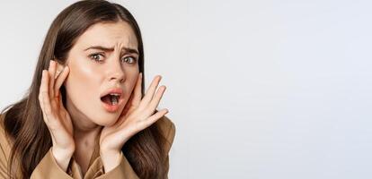 Image of sad girl looking in mirror and touching facial skin, shocked and frustrated over bad skin condition, standing over white background photo