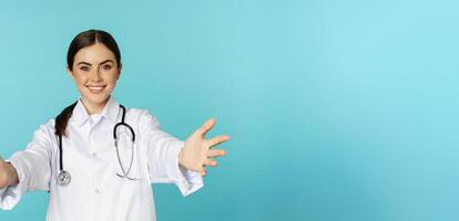 Smiling friendly doctor, girl healthcare worker, intern reaching hands, inviting, hugging or receiving in arms, standing over torquoise background photo