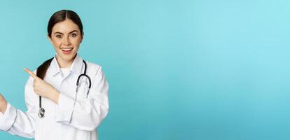 Portrait of smiling medical worker, girl doctor in white coat with stethoscope, pointing fingers left, showing medical clinic advertisement, torquoise background photo