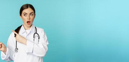 Surprised female doctor, physician with stethoscope, pointing and looking left with amazed, wow face, standing over torquoise background photo