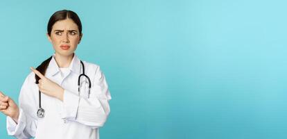 Confused, disappointed medical worker, frowning while pointing fingers left with doubtful, puzzled face expression, standing over torquoise background photo