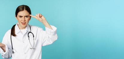 Annoyed woman doctor pointing finger at head, scolding someone stupid, crazy or strange, standing over torquoise background photo