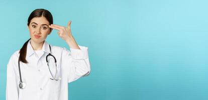 Tired and annoyed young woman showing pictol shot gesture over head, looking upset or disappointed, standing over torquoise background photo
