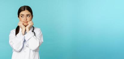 Scared and worried woman doctor in white coat, looking anxious and insecure, shaking from fear, standing over torquoise background photo