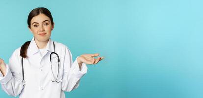 Portrait of doctor, woman intern shrugging shoulders, smiling and saying sorry, dont know, being clueless, standing over blue background photo