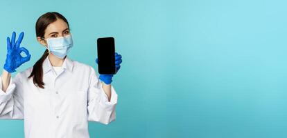 Portrait of doctor in medical face mask and gloves, showing mobile phone app, smartphone screen and okay sign, recommending online checkup website, standing over torquoise background photo
