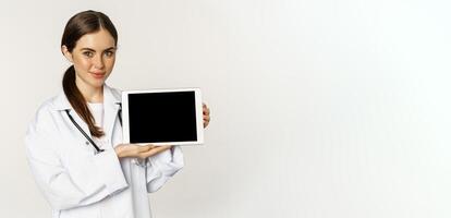 Online medical help, remote appointment. Smiling beautiful woman doctor showing digital tablet screen, demonstrating website logo, standing over white background photo