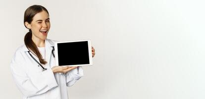 Online medical help, remote appointment. Smiling beautiful woman doctor showing digital tablet screen, demonstrating website logo, standing over white background photo