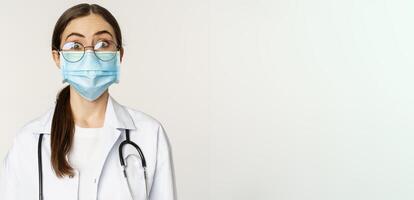 Medical worker, young doctor looking amazed and surprised, wearing face mask to prevent catching covid-19, standing over white background photo