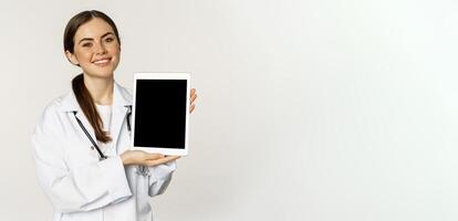 Online medical help, remote appointment. Smiling beautiful woman doctor showing digital tablet screen, demonstrating website logo, standing over white background photo