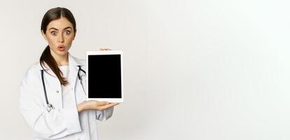 Image of woman doctor, female healthcare worker showing online medical website, digital tablet screen and smiling, standing in white coat over white background photo