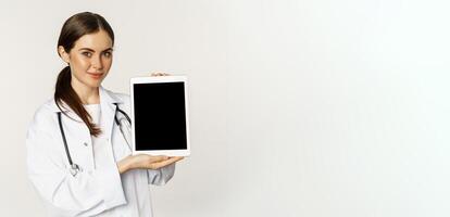 Image of woman doctor, female healthcare worker showing online medical website, digital tablet screen and smiling, standing in white coat over white background photo