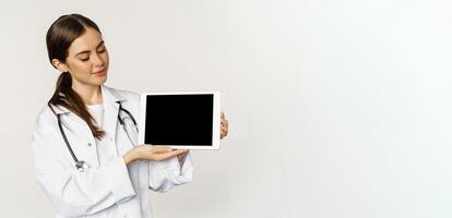 Image of woman doctor, female healthcare worker showing online medical website, digital tablet screen and smiling, standing in white coat over white background photo