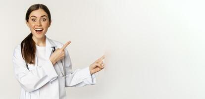 Woman doctor, physician in medical uniform, pointing fingers right, showing banner advertisement, smiling amazed, checking out promo discount, white background photo