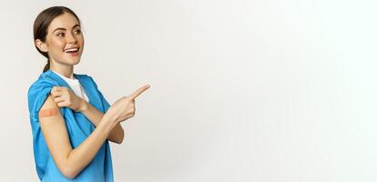 Smiling nurse, female healthcare worker in scrubs, pointing finger right, showing patch on shoulder, vaccination campaign, covid-19 and pandemic concept, white background photo