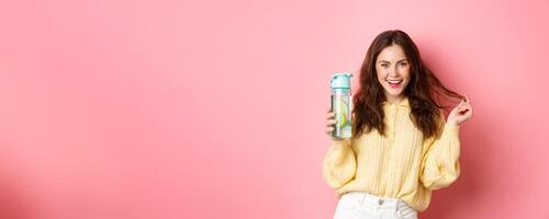Sassy attractive woman looking confident at camera, holding personal bottle with lemon water healthy drink, standing against pink background photo