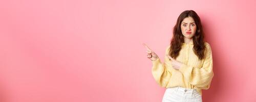 Sad and gloomy brunette girl complaining on bad promotional text, pointing fingers aside to left, showing banner with disappointed expression, standing over pink background photo