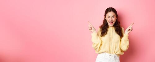 Image of cheerful good-looking female model pointing fingers aside, showing two choices sidewas, left and right logo, standing over pink background photo