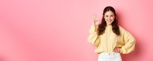 Portrait of young confident woman smiling pleased, say yes while showing okay sign, give approval, praise excellent choice, standing against pink background photo