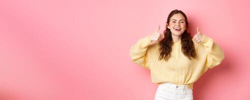 Very good, well done. Smiling girl being supportive, laughing and showing thumbs up in approval, like awesome idea, praise you, standing against pink background photo