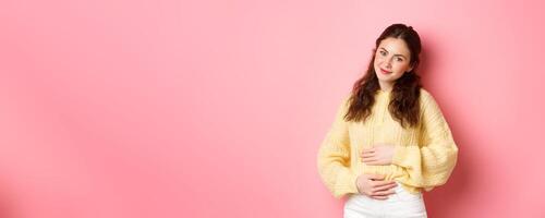 Young smiling woman being pregnant, rubbing her belly and looking with hopeful happy face at camera, expecting for baby, standing against pink background photo