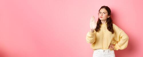 Angry and bossy young woman frowning, looking serious, showing block stop gesture, stretch out hand to say no, refuse something bad, forbid action, standing against pink background photo