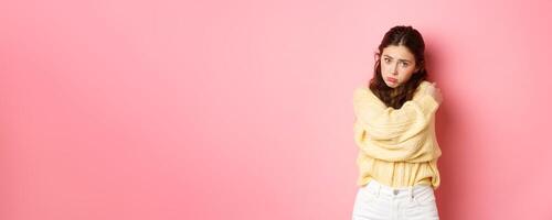 Cute silly girl making sad face and looking with puppy eyes at camera, hugging herself, standing innocent against pink background photo