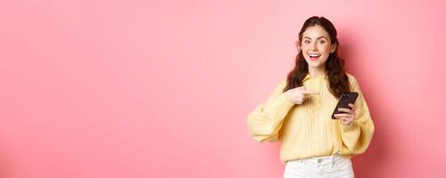 Young attractive woman holding smartphone, pointing at screen, promoting, talking about her social media page, standing over pink background photo