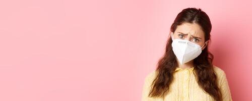 Preventive measures, health care concept. Close up of sad young woman in medical respirator express compassion, look with pity and upset face, standing against pink background photo