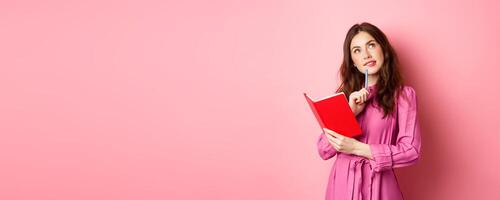 Beautiful young woman look thoughtful, writing in notebook, holding planner or diary, plan her schedule, standing against pink background photo