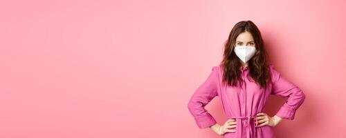 Covid-19, pandemic and lifestyle concept. Angry woman in medical respirator look with judgement and dislike, frowning and scolding someone without face mask, pink background photo