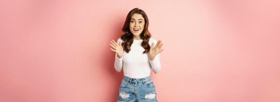 Image of excited smiling stylish woman, shaking hands and jumping enthusiastic, looking amazed at camera, standing against pink background photo