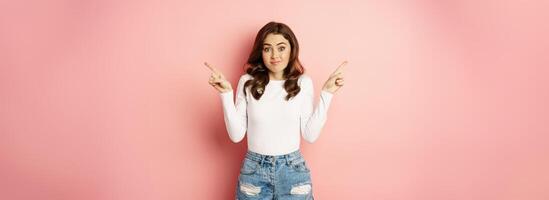 Glamour indecisive girlfriend, shrugging and pointing fingers sideways, showing left and right promo text, store offers, confused what to choose, pink background photo