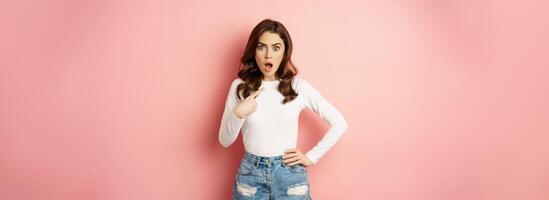 Shocked and offended young beautiful woman, pointing finger at herself, oh my gosh, who me gesture, being insulted, standing in disbelief over pink background photo