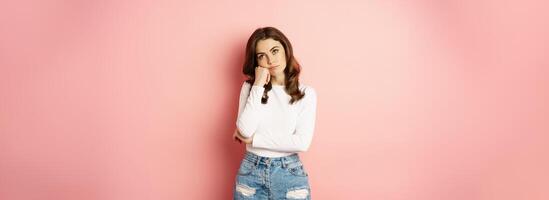 Image of sad and bored, reluctant young woman looking at smth boring, feeling lonely, leaning head on fist with indifferent face expression, standing over pink background photo