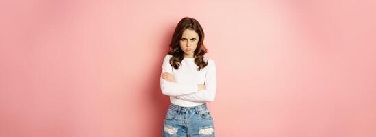 Angry silly girl cross arms offended, looking from under forehead and frowning disappointed, insulted by someone, standing pissed off over pink background photo