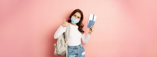 Covid and travelling concept. Happy woman traveller in medical mask, holding passport and airplane tickets, going on trip, vacation during quarantine, pink background photo