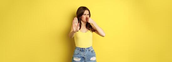 Woman showing stop sign and shut nose from disgust, dislike bad smell, declining something awful, standing over yellow background photo