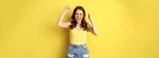 Enthusiastic young woman shouting and cheering, rejoicing, raising hands up and screaming in joy, chanting, standing over yellow background photo