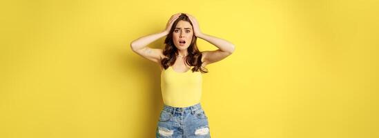 Troubled and shocked woman holding hands on head, panicking, feeling distressed and anxious, having problem, standing over yellow background photo