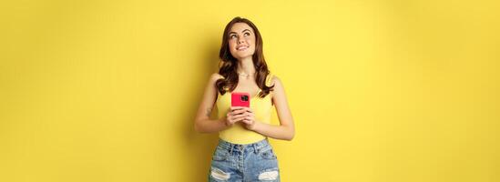 Technology and cellphone. Beautiful stylish girl order in online store, praying in application, using smartphone app and thinking, making decision, standing over yellow background photo