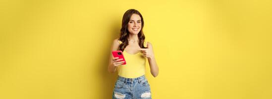 Cellular technology and online shopping concept. Beautiful smiling woman pointing finger at her mobile phone, recommending website, app on smartphone photo
