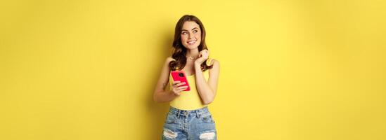 Technology and cellphone. Beautiful stylish girl order in online store, praying in application, using smartphone app and thinking, making decision, standing over yellow background photo