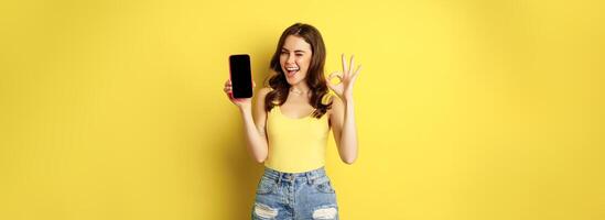 Pretty brunette woman, showing mobile phone screen, smartphone app interface and okay sign, recommend website, store, standing against yellow background photo