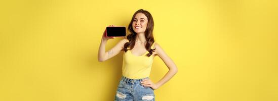 Proud smiling woman showing mobile phone horizontal screen, looking proud and satisfied, demonstrating smartphone display, app or online shopping store, yellow background photo