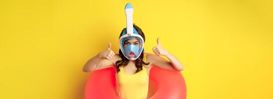 Funny happy woman in swimming ring, wearing snorkling mask for diving, showing thumbs up, good approval gesture, posing against yellow background photo