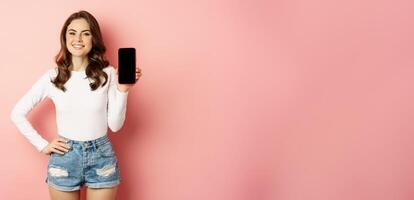 Stylish sexy woman showing mobile phone screen, smartphone app interface and smiling, demonstarting online shop, standing over pink background photo