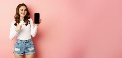 Proud and happy smiling girl pointing finger, showing mobile phone screen, cellphone app or promo text on smartphone, standing over pink background photo