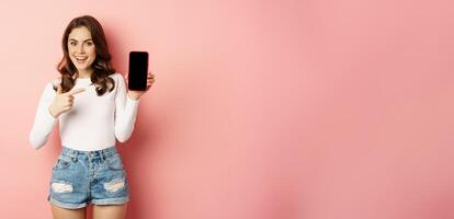 Stylish sexy woman showing mobile phone screen, smartphone app interface and smiling, demonstarting online shop, standing over pink background photo
