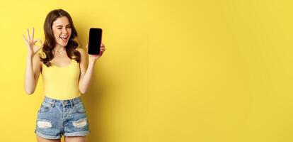 Pretty brunette woman, showing mobile phone screen, smartphone app interface and okay sign, recommend website, store, standing against yellow background photo